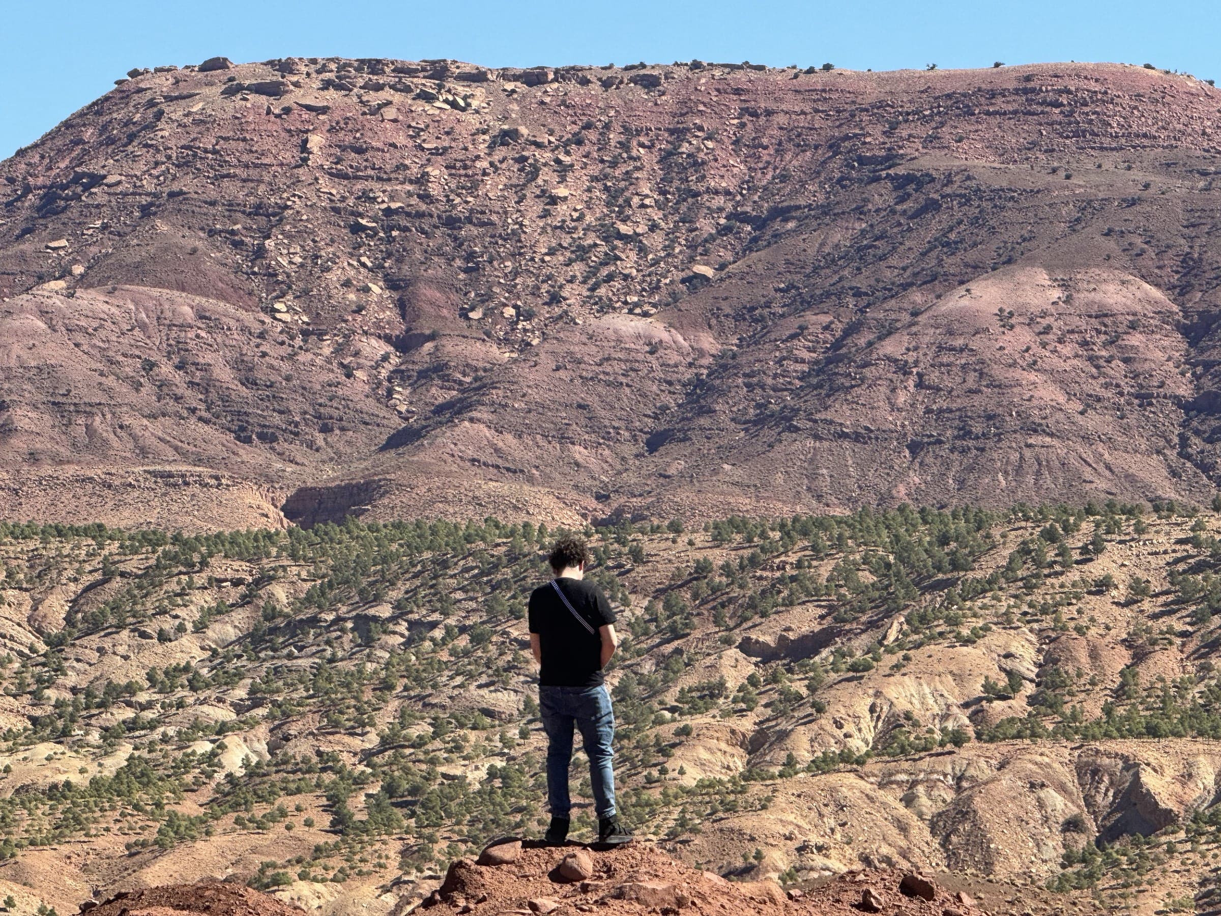 Viajero contemplando el paisaje árido y montañoso del Atlas marroquí con vegetación de enebros y cielo azul