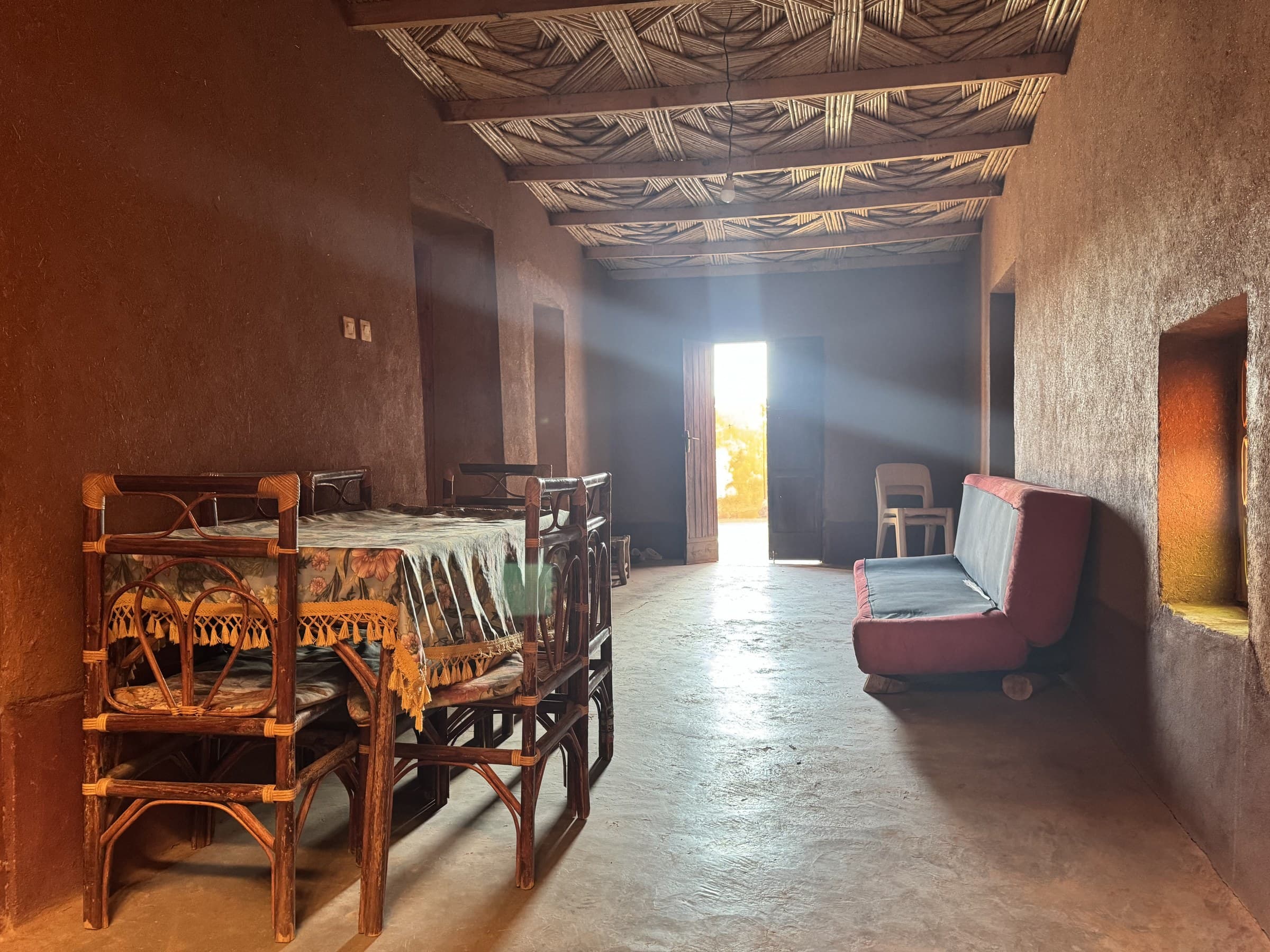 Interior de casa bereber de adobe con mesa de caña, sofá y rayos de luz entrando por la puerta en el desierto de Marruecos