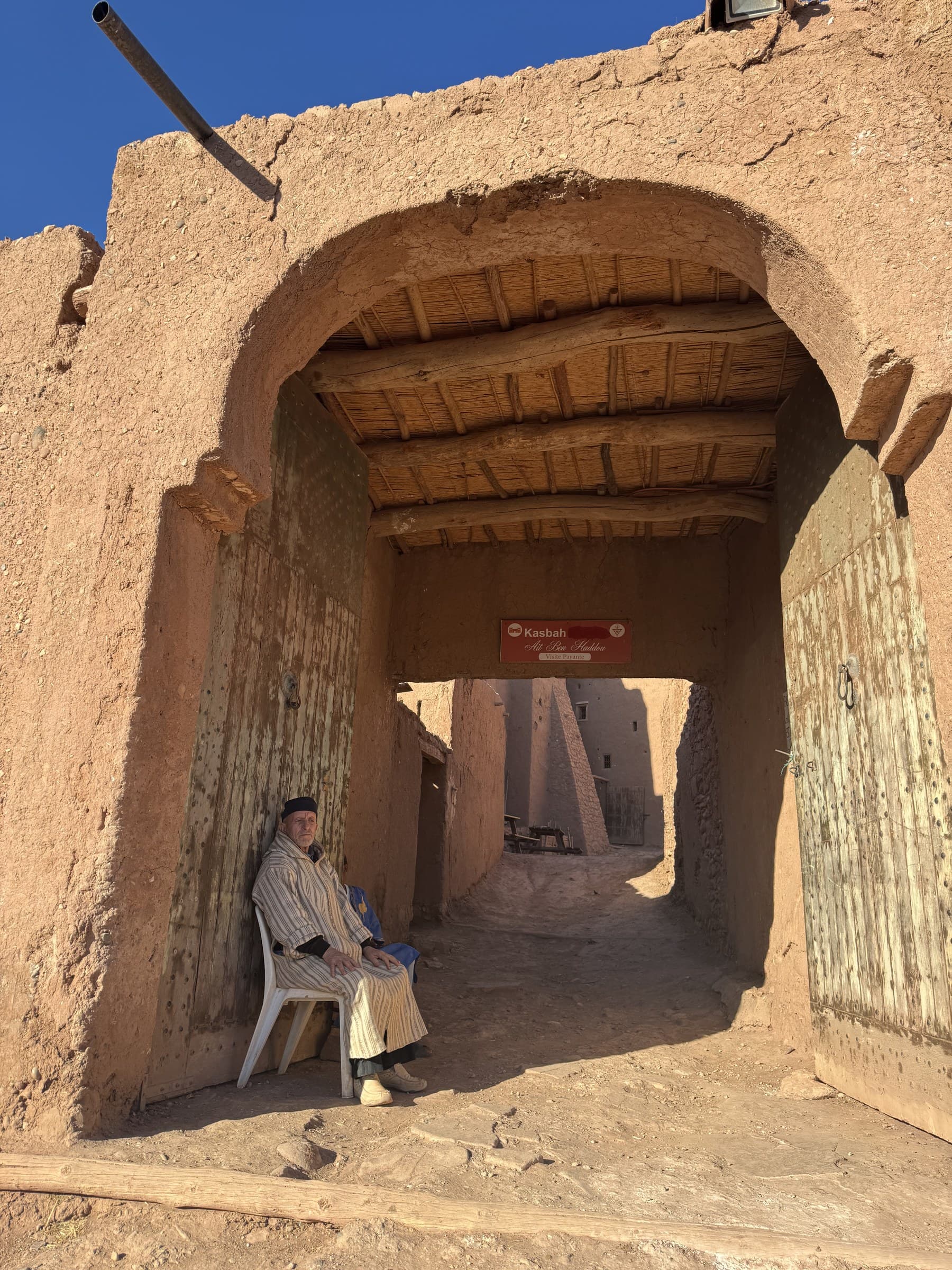 Entrada de la Kasbah Aït Ben Haddou con hombre bereber sentado bajo arco de adobe, Patrimonio UNESCO Marruecos