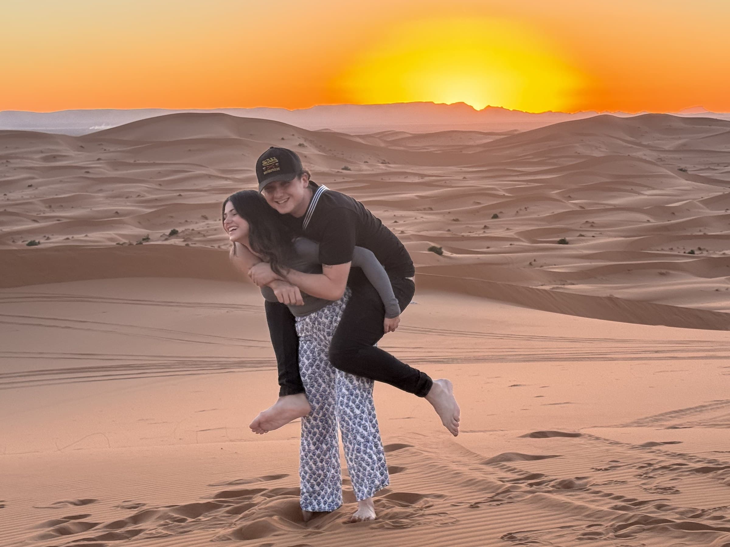 Pareja jugando y riendo sobre las dunas del desierto de Merzouga al atardecer con cielo naranja