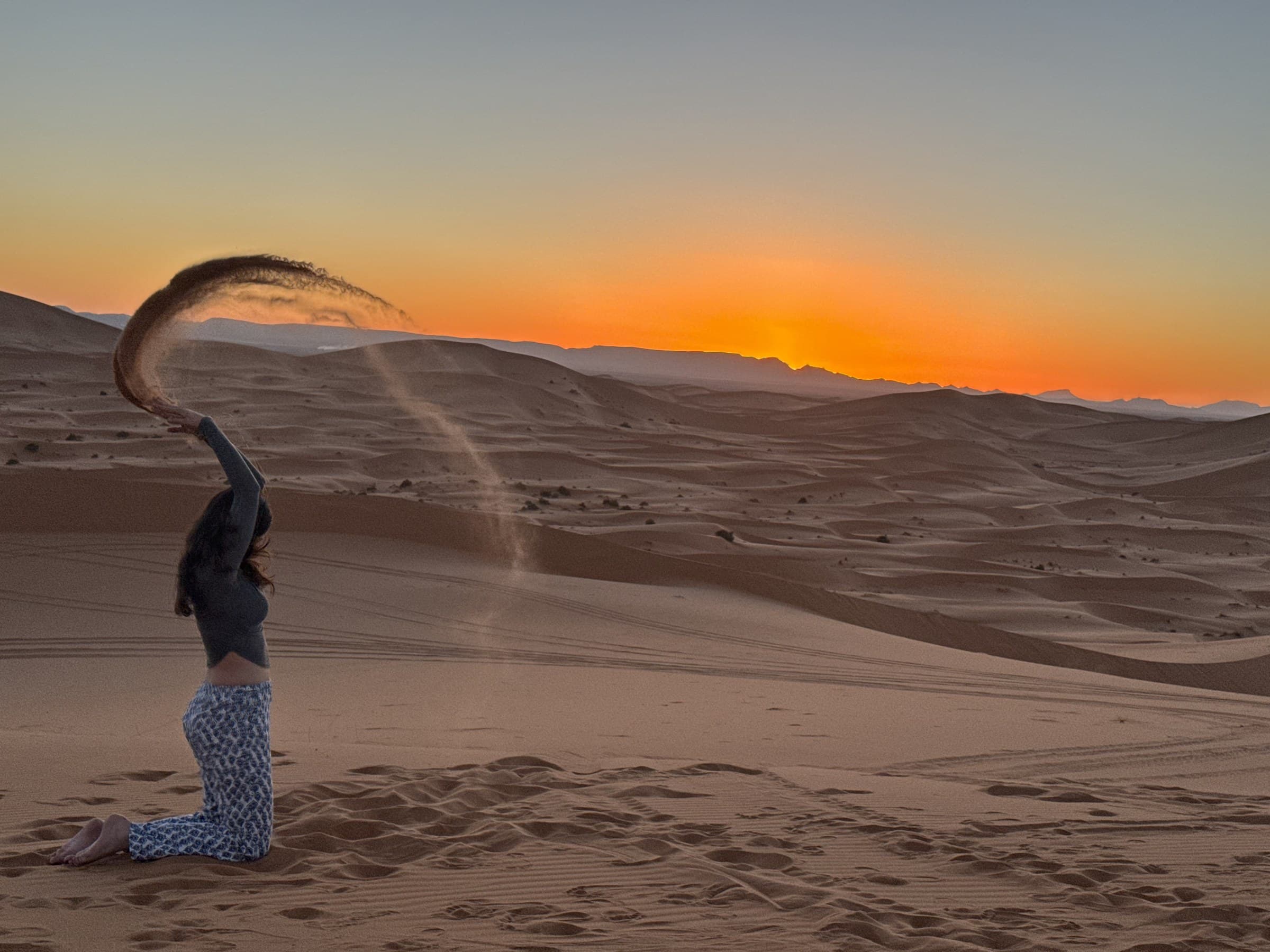 Mujer lanzando arena al atardecer en las dunas del Sahara de Merzouga, cielo naranja intenso