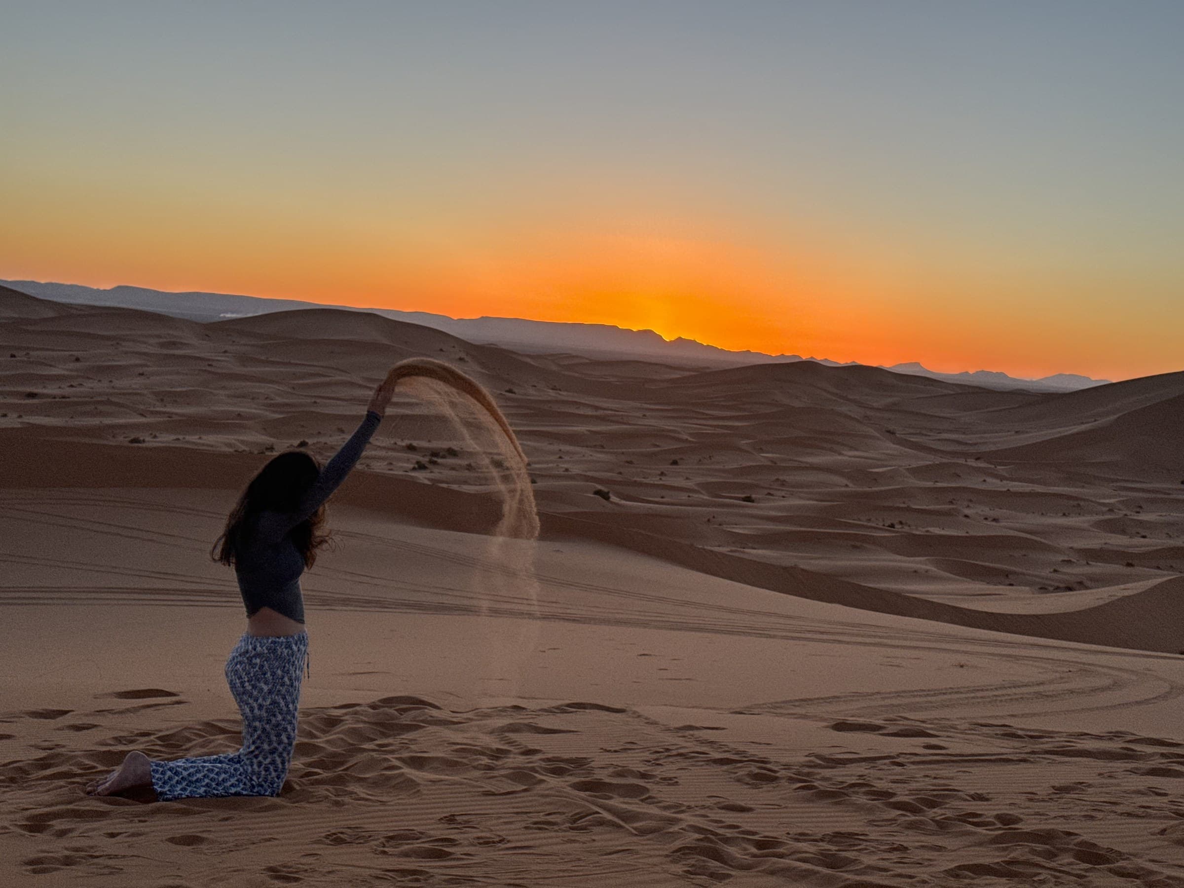 Mujer lanzando arena en las dunas del Erg Chebbi al atardecer con cielo naranja en Merzouga, Marruecos