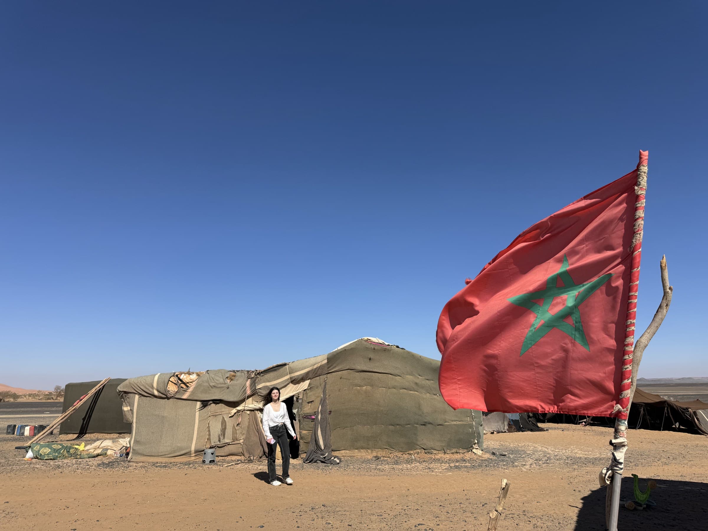 Viajera junto a jaima nómada en el desierto de Merzouga con la bandera de Marruecos ondeando bajo cielo azul