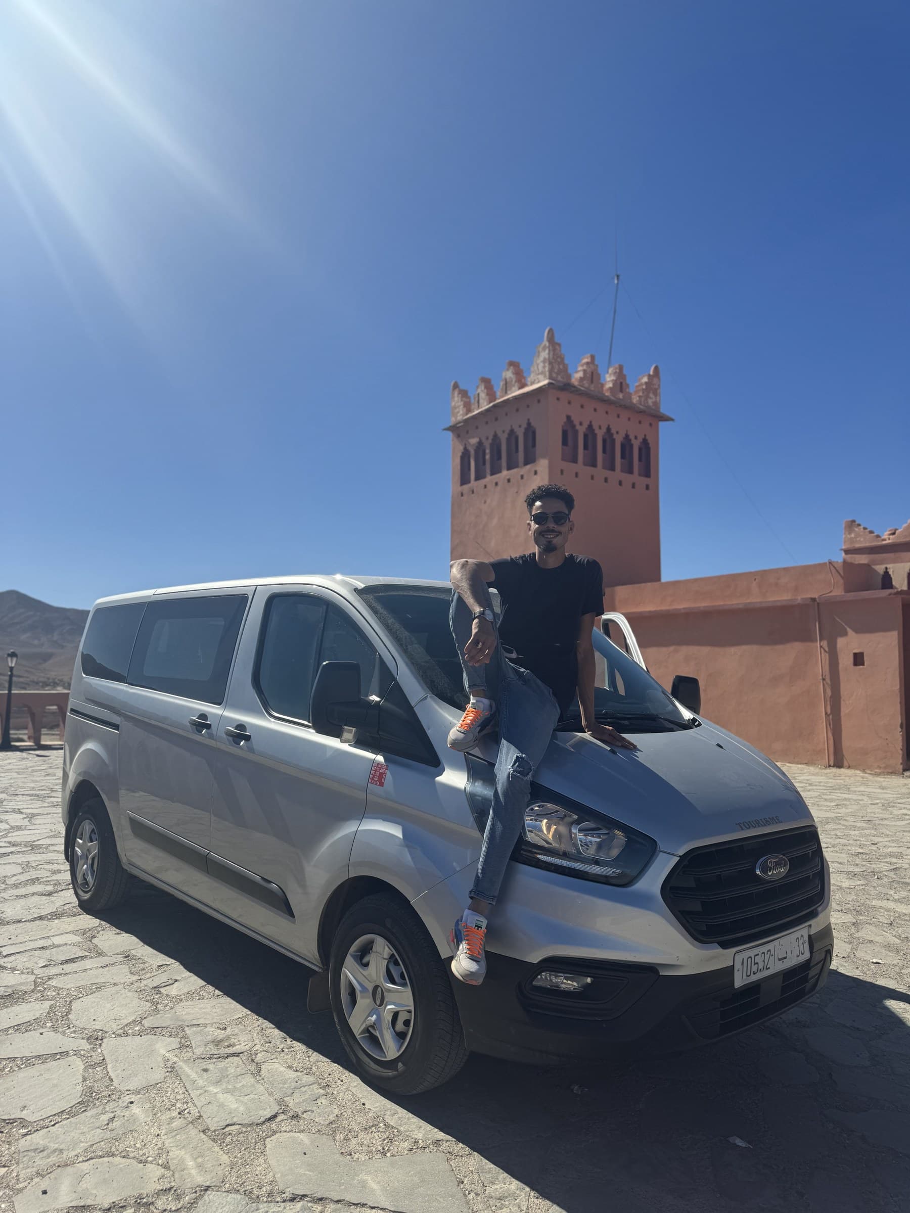 Guía privado apoyado en furgoneta Ford Transit frente a torre de kasbah de barro en Ouarzazate, Marruecos
