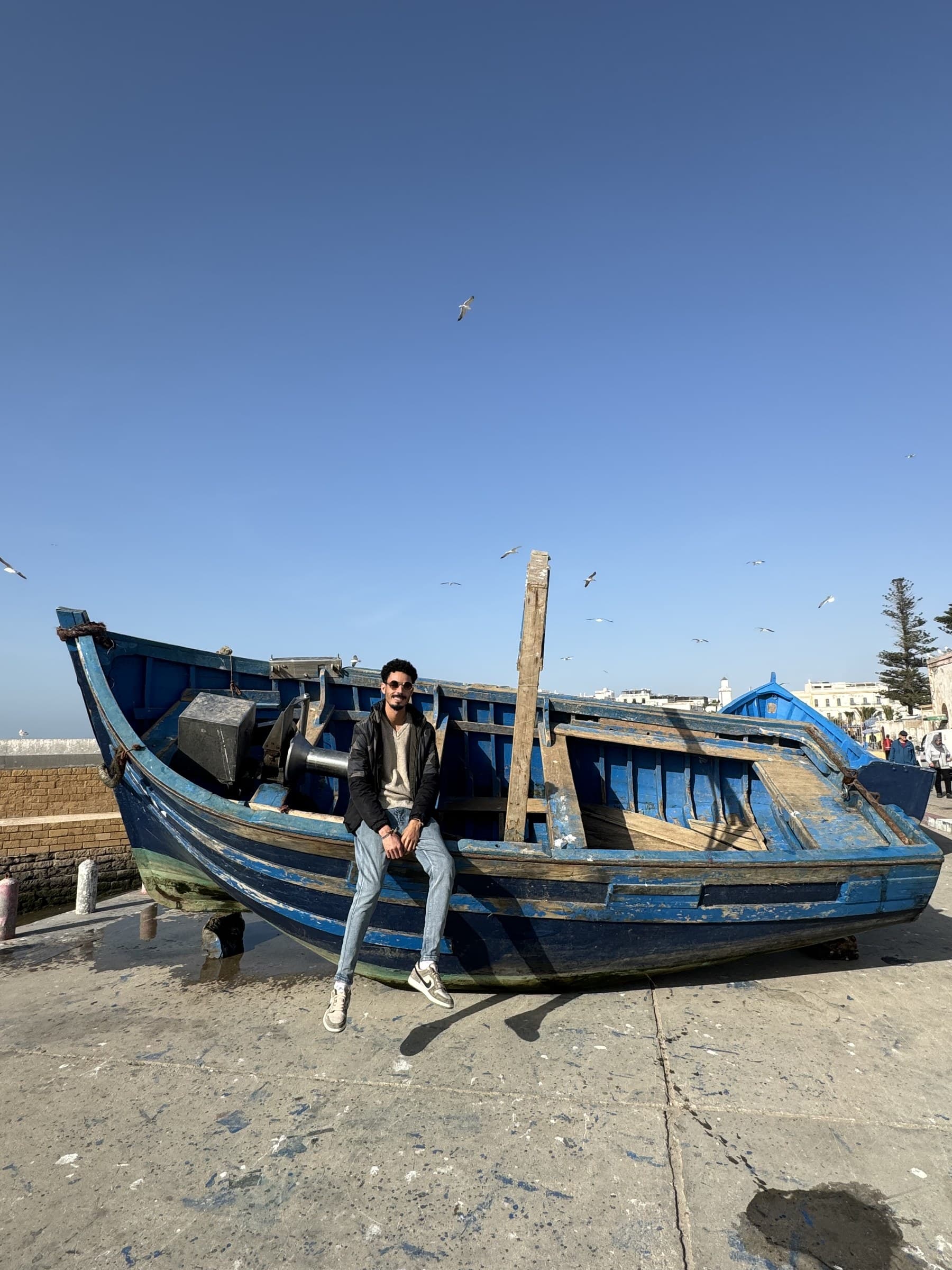 Hombre apoyado en barca de pesca azul tradicional en el puerto de Essaouira con gaviotas volando