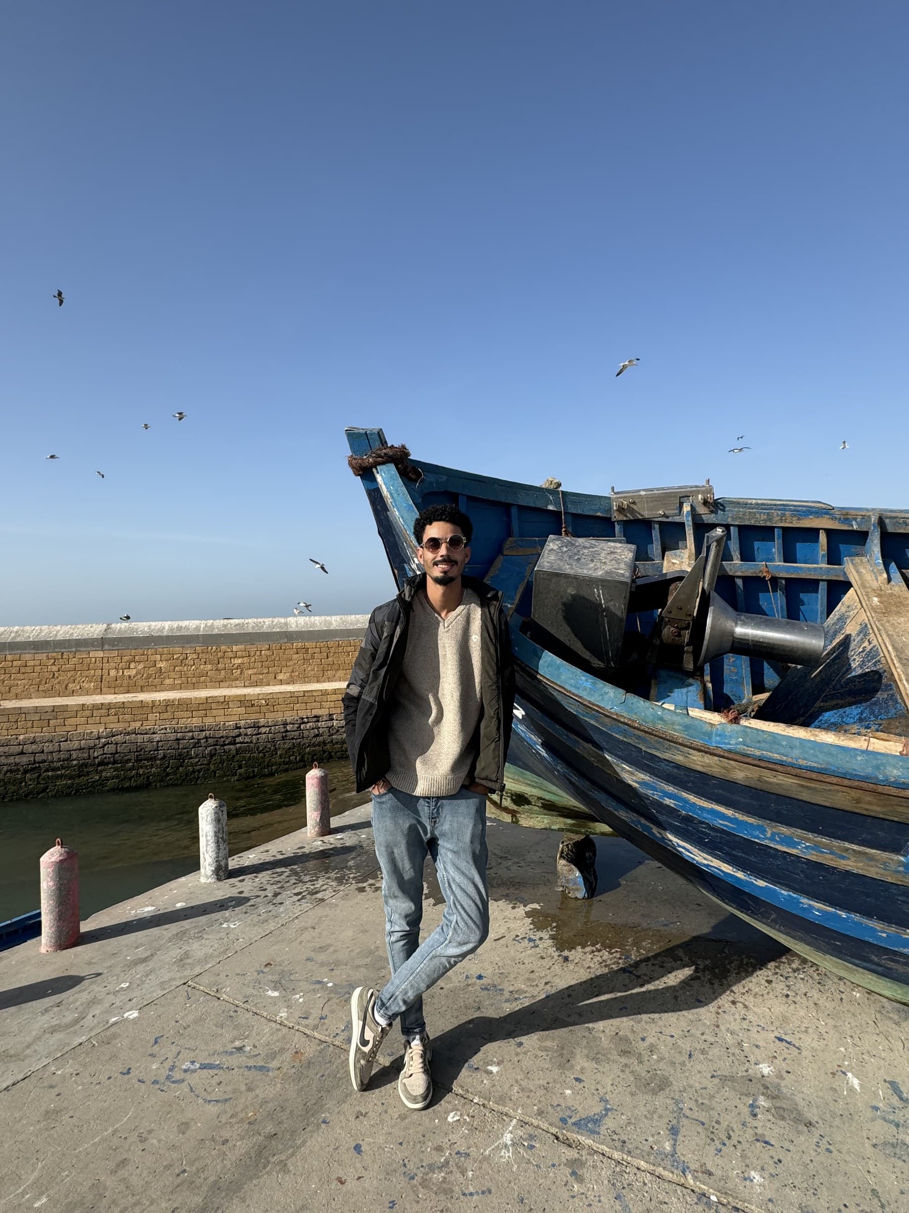 Joven posando junto a barco de pesca azul en el puerto de Essaouira con gaviotas volando al fondo
