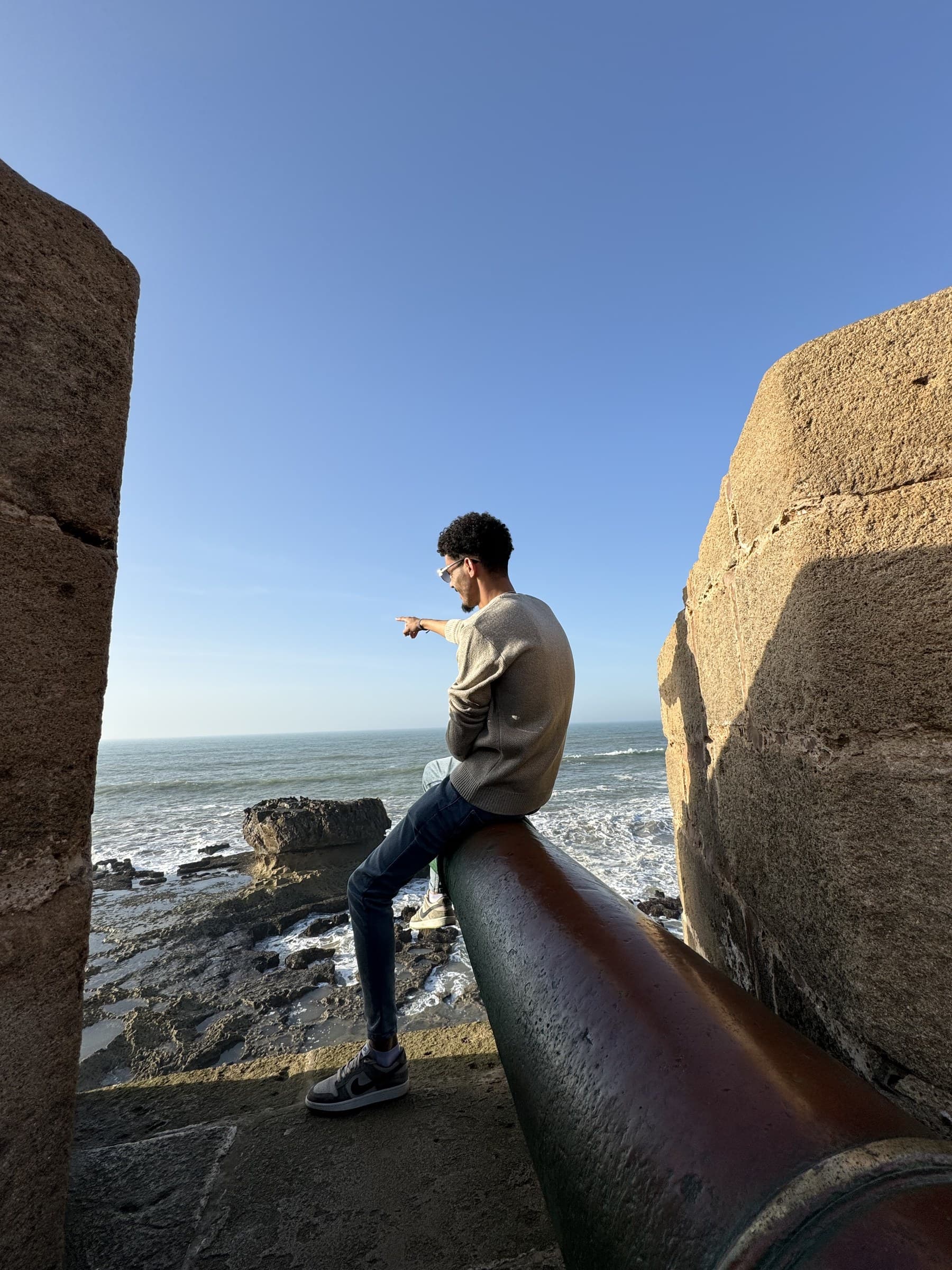 Joven sentado en un cañón histórico señalando el océano Atlántico desde las murallas de Essaouira, Marruecos