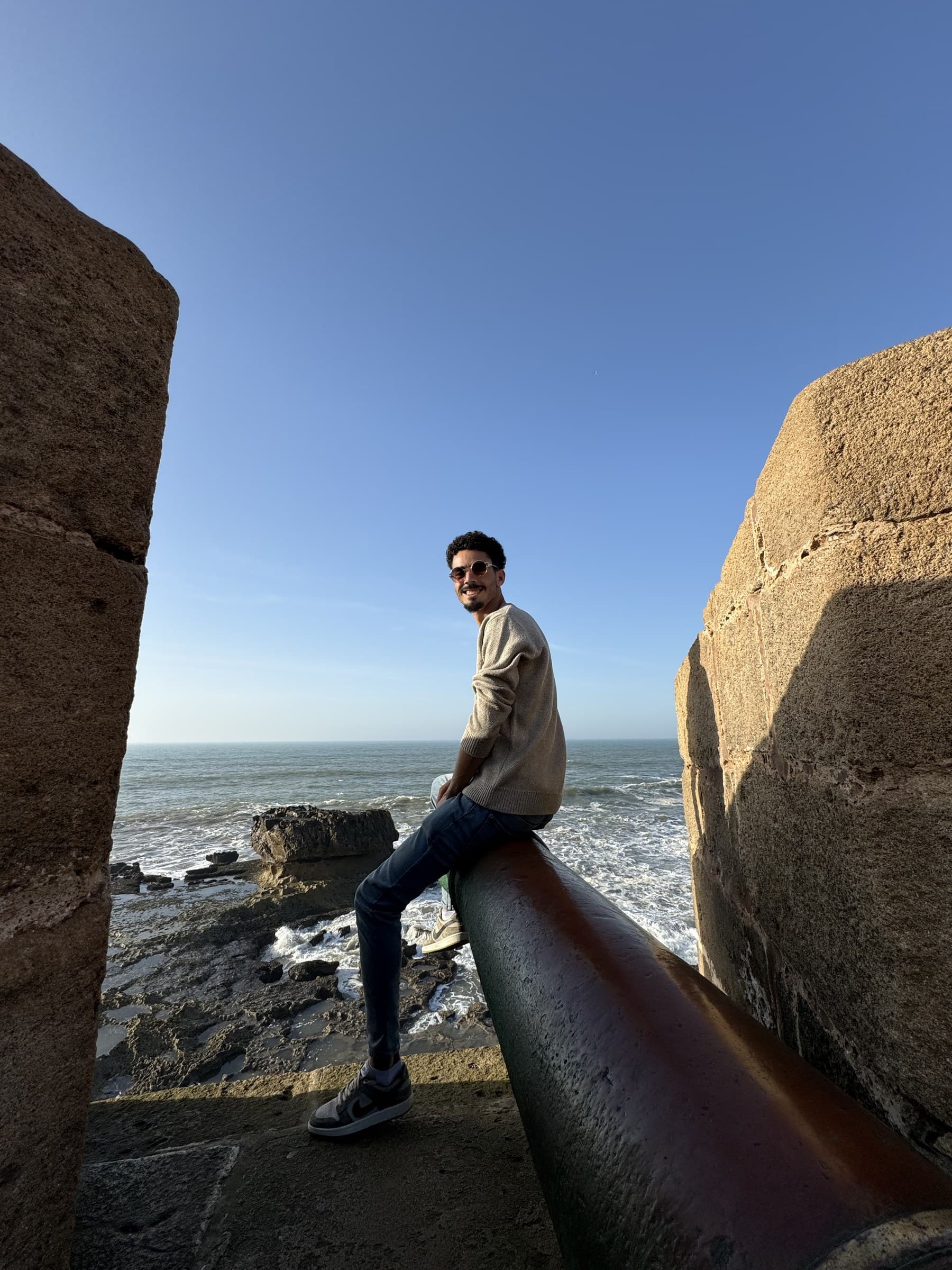 Joven sentado en cañón antiguo frente al océano Atlántico en las murallas de Essaouira, Marruecos