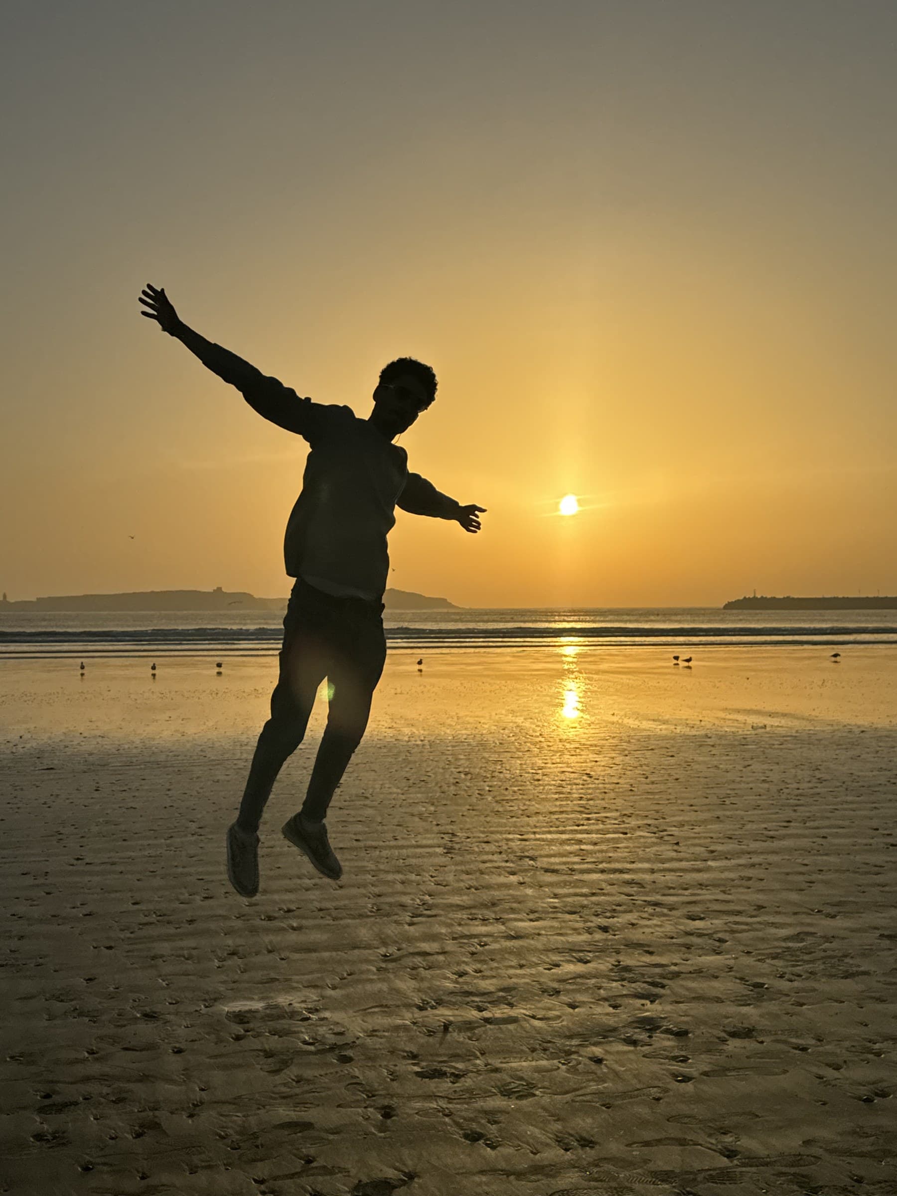 Silueta de persona saltando en la playa al atardecer en Essaouira, Marruecos, con el sol reflejado en el agua