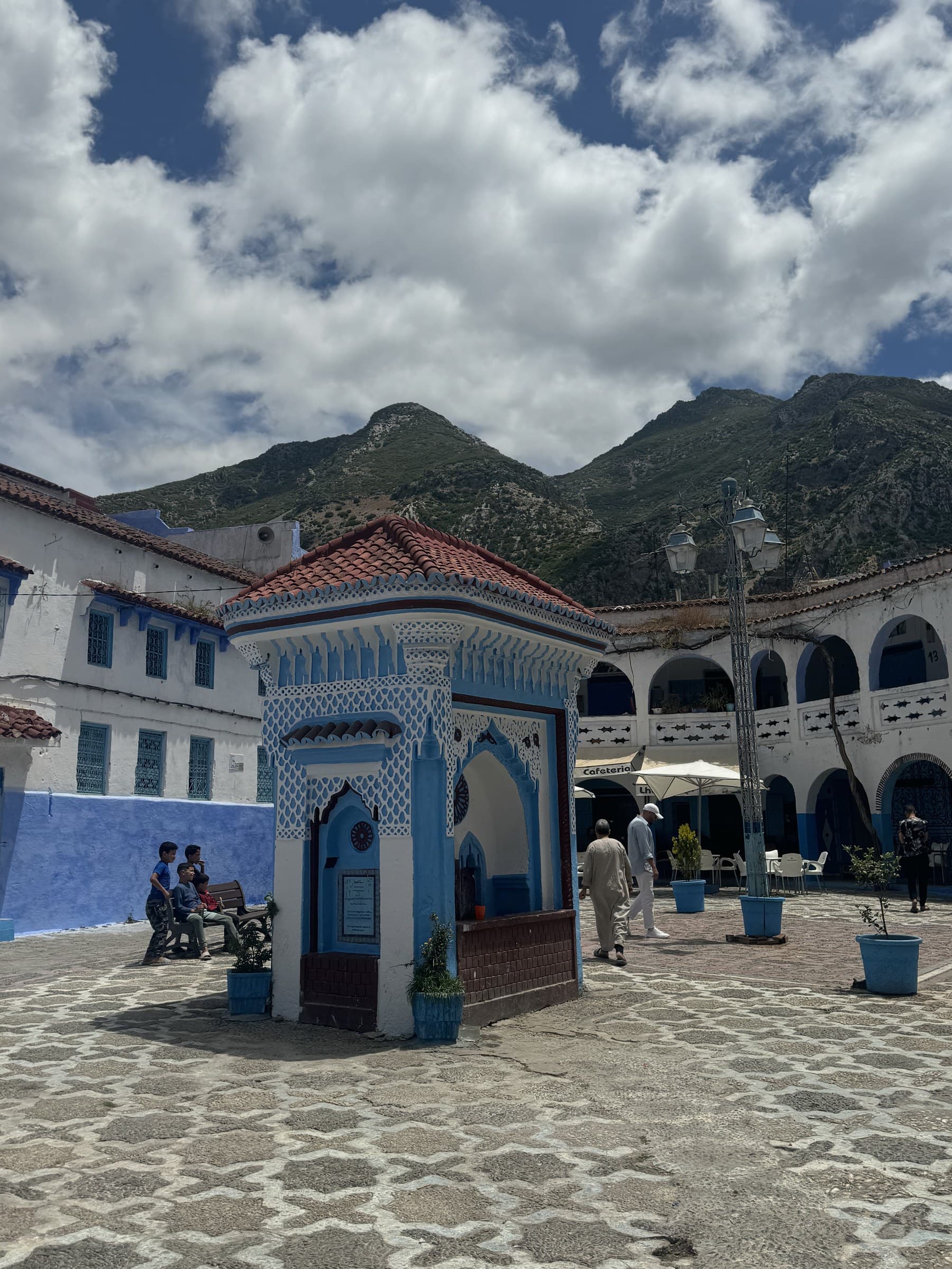 Plaza Uta el-Hammam de Chefchaouen con kiosco azul y blanco, arcos y montañas del Rif al fondo