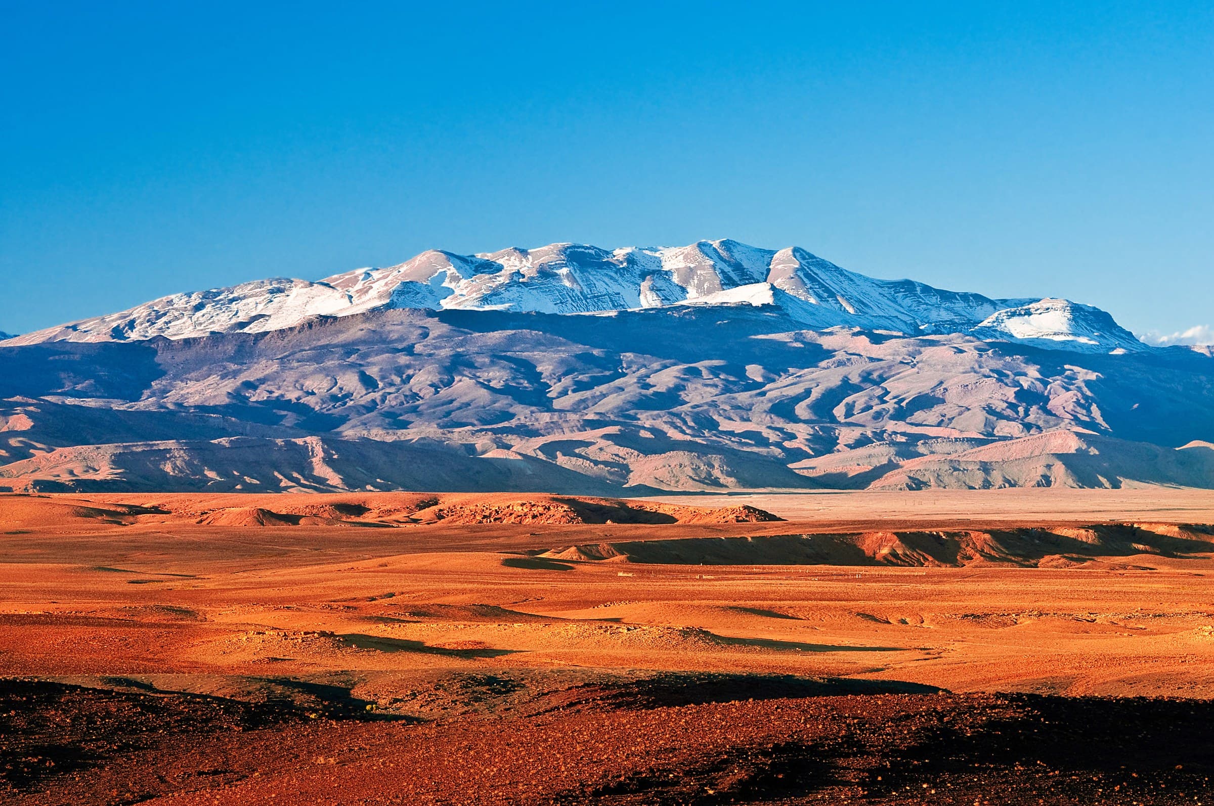 Paisaje del Alto Atlas marroquí con cumbres nevadas sobre llanura rocosa rojiza bajo cielo azul intenso